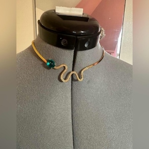 May include: A gold-coloured choker necklace with a wavy design and a large, round, teal-coloured gemstone. The necklace is displayed on a grey mannequin. The wavy design is embellished with small, clear crystals.