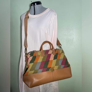 May include: A brown and multicoloured satchel bag with a geometric pattern on the top half and a solid brown leather bottom. The bag has a top handle and a detachable shoulder strap. The bag is displayed on a mannequin.
