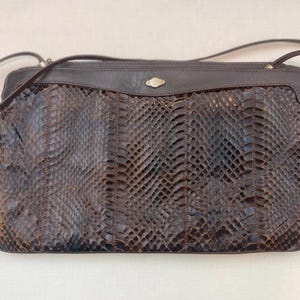 May include: A brown leather crossbody bag with a snakeskin pattern. The bag has a rectangular shape, a flap closure, and a long, thin strap. The bag is dark brown with a silver-coloured clasp.