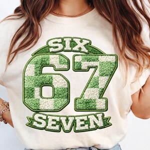 May include: Cream-colored t-shirt featuring the text "SIX 67 SEVEN" in a green and white checkered pattern. The numbers and words have a textured, fuzzy appearance, with a green border. The shirt is a casual, everyday wear item.