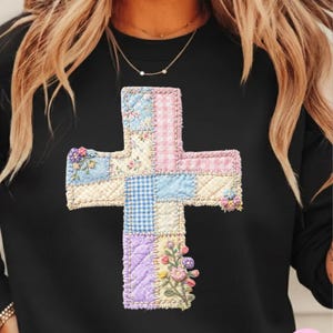 May include: Black sweatshirt featuring a patchwork cross design. The cross is composed of various fabric patterns, including floral, gingham, and solid colors in pastel shades of blue, pink, and purple. The cross is embellished with floral accents.