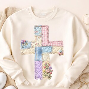 May include: Cream-colored sweatshirt featuring a patchwork cross design. The cross is composed of various pastel-colored fabrics, including floral, gingham, and solid patterns. Embroidered floral accents are placed on the cross.