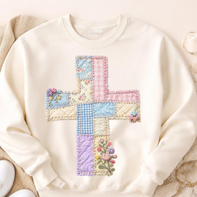 May include: Cream-colored sweatshirt featuring a patchwork cross design. The cross is composed of various pastel-colored fabrics, including floral, gingham, and solid patterns. Embroidered floral accents are placed on the cross.