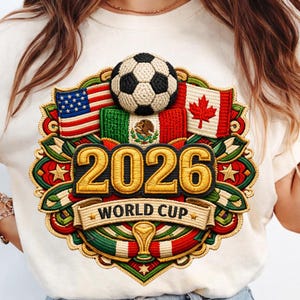 Peut inclure: T-shirt crème avec un motif brodé pour la Coupe du Monde 2026. Le motif comprend un ballon de football, les drapeaux des États-Unis, du Mexique et du Canada, et le texte "2026 WORLD CUP".
