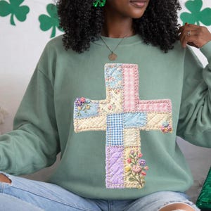May include: Sage green sweatshirt featuring a patchwork cross design. The cross is made of various fabric patterns, including floral, gingham, and solid colors, with embroidered floral accents. The sweatshirt has a crew neck and long sleeves.