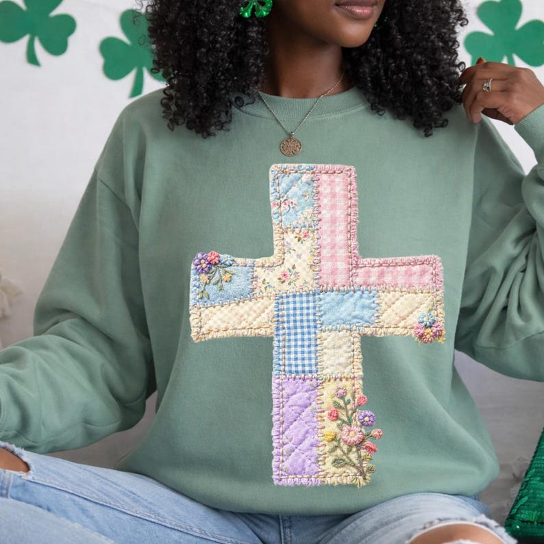 May include: Sage green sweatshirt featuring a patchwork cross design. The cross is made of various fabric patterns, including floral, gingham, and solid colors, with embroidered floral accents. The sweatshirt has a crew neck and long sleeves.