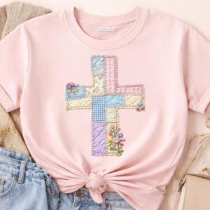 May include: A light pink t-shirt featuring a cross design. The cross is made of patchwork squares in pastel colors, including blue, pink, and yellow, with floral and gingham patterns. The shirt has short sleeves and a tied knot at the waist.