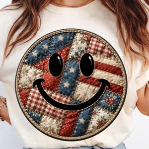 May include: Cream-colored t-shirt featuring a large, circular smiley face design. The smiley face is a patchwork of red, white, and blue fabrics, resembling the American flag. The design includes stars, stripes, and floral patterns, with a black outline and eyes.
