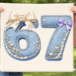 May include: A canvas featuring the numbers 67, crafted from denim-like fabric. The numbers are embellished with pearls, floral accents, and bows. The number 6 has a cream-colored bow, while the 7 has a purple bow.
