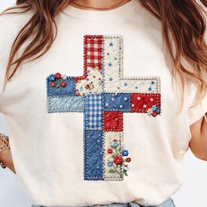 Puede incluir: Camiseta color crema con un diseño de cruz hecho de tela de retazos. La cruz incorpora patrones rojos, blancos y azules, incluyendo cuadros, flores y telas con estrellas. Bordados florales acentúan la cruz.