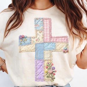 May include: Cream-colored t-shirt featuring a patchwork cross design. The cross is composed of various fabric patterns, including floral, gingham, and solid colors in pastel shades. Embroidered flowers accent the cross. A casual and comfortable top.