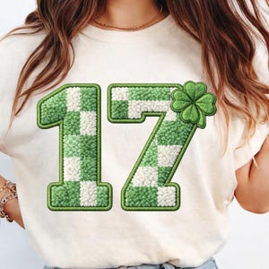 May include: Cream-colored t-shirt featuring a large, green and white checkered "17" design with a shamrock accent. The numbers have a textured, fuzzy appearance, and the shamrock is a bright green color. The shirt is likely for St. Patrick's Day.