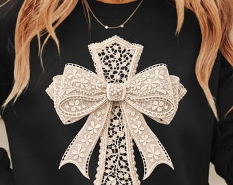 May include: Black long-sleeve shirt featuring a cream-colored, lace-like cross design. The cross has a bow at the center and floral details. The shirt is made of a soft material.
