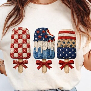 May include: White t-shirt featuring three patriotic popsicle designs. The popsicles have red, white, and blue patterns, including checks, stars, stripes, and polka dots. Each popsicle has a wooden stick and a red bow.