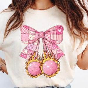 Könnte beinhalten: Cremefarbenes T-Shirt mit einem rosa Schleifenmuster im Patchwork-Stil und Herzakzenten. Unter der Schleife befinden sich zwei rosa Kugeln in Flammen. Das Grafik-T-Shirt hat ein skurriles und einzigartiges Design.