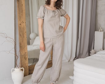 Puede incluir: Un conjunto de pijama de lino beige claro. La parte superior tiene mangas cortas y un lazo en el escote. Los pantalones son holgados con tobillos fruncidos. El conjunto está hecho de una tela suave y transpirable, perfecto para descansar o dormir.