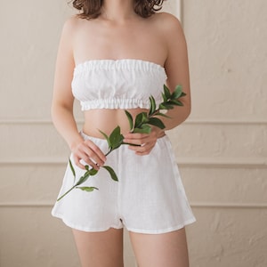 May include: A white linen two-piece set, featuring a strapless crop top and matching shorts. The top has a ruffled edge, and the shorts are high-waisted. The model is holding a green leafy branch. The background is a neutral beige.