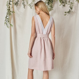 Pode incluir: Um vestido rosa claro sem mangas com decote em V e acabamento em renda branca. O vestido apresenta um cinto a condizer e vai até aos joelhos. A modelo usa saltos bege. O fundo é um tecido neutro com folhagem verde.