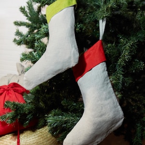 Pode incluir: Duas meias de Natal com corpos cinza claro e punhos coloridos penduradas em uma árvore de Natal. Uma meia tem uma punho verde e a outra um punho vermelho. Ambas têm laços brancos para pendurar.