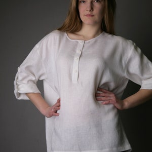 May include: A white linen shirt with a button-up placket and rolled-up sleeves. The shirt is loose-fitting and has a relaxed silhouette.