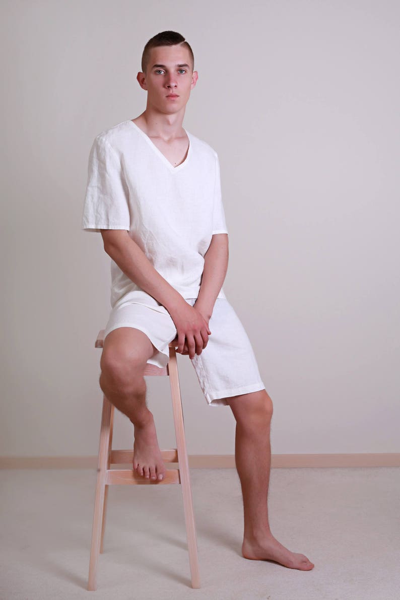 Pode incluir: Uma pessoa jovem vestindo uma camisa e shorts de linho branco senta-se em um banquinho de madeira. A pessoa tem cabelo castanho curto e est&aacute; olhando diretamente para a c&acirc;mera.