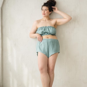 May include: A sage green two-piece set consisting of a strapless bandeau top with a gathered elastic neckline and matching high-cut shorts with an elastic waistband. The garment features a soft, textured fabric finish in a muted seafoam tone.