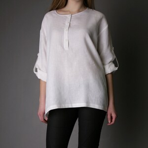 May include: A white linen shirt with a button-up placket and rolled-up sleeves. The shirt is loose-fitting and has a relaxed silhouette. The model is wearing black leggings.