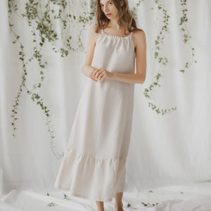 Puede incluir: Un vestido largo de lino beige claro con tirantes finos y un dobladillo con volantes. El vestido es largo hasta el suelo y tiene un diseño sencillo y elegante. La modelo lleva bailarinas rosa claro. El fondo es una cortina blanca con vides verdes.