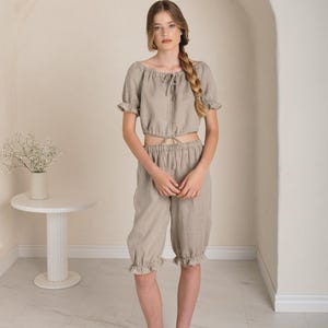 May include: A woman is wearing a light brown linen top and matching shorts. The top has a square neckline and a tie at the front. The shorts have a ruffled hem. She is wearing white flats.