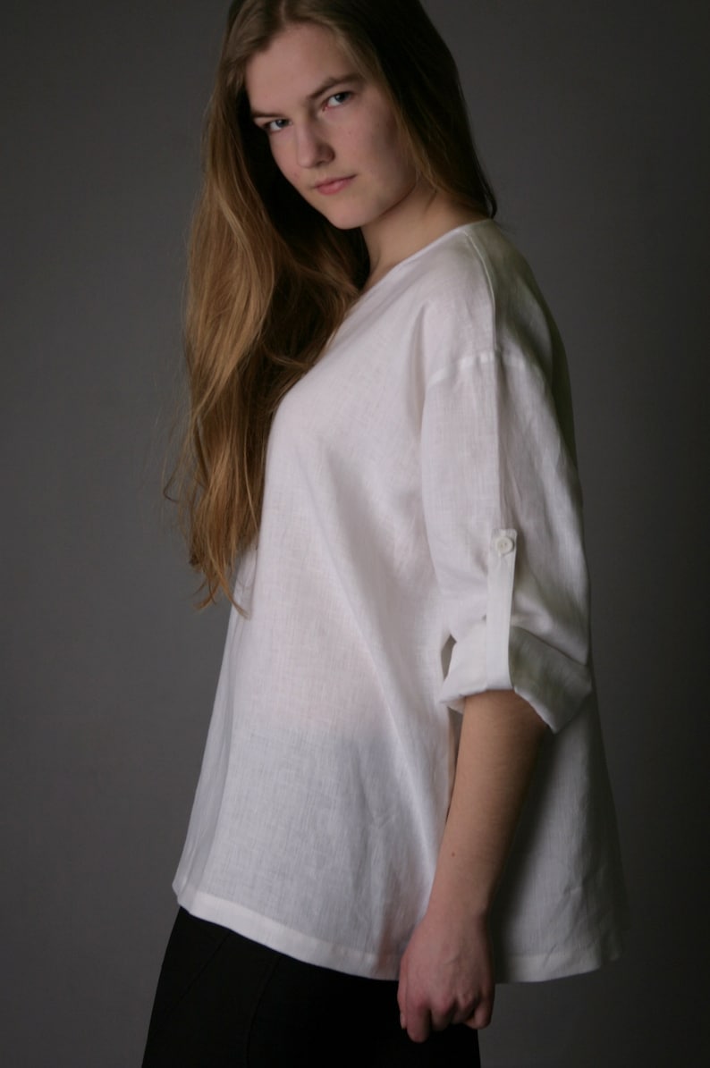 May include: A white linen shirt with a relaxed fit and rolled up sleeves. The shirt has a v-neck and a button on the sleeve.