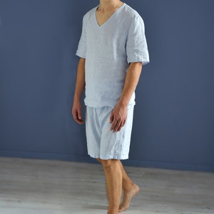 Pode incluir: Um homem vestindo uma camisa de linho azul claro e shorts combinando. Ele est&aacute; em p&eacute; em um piso de madeira clara em frente a uma parede azul.