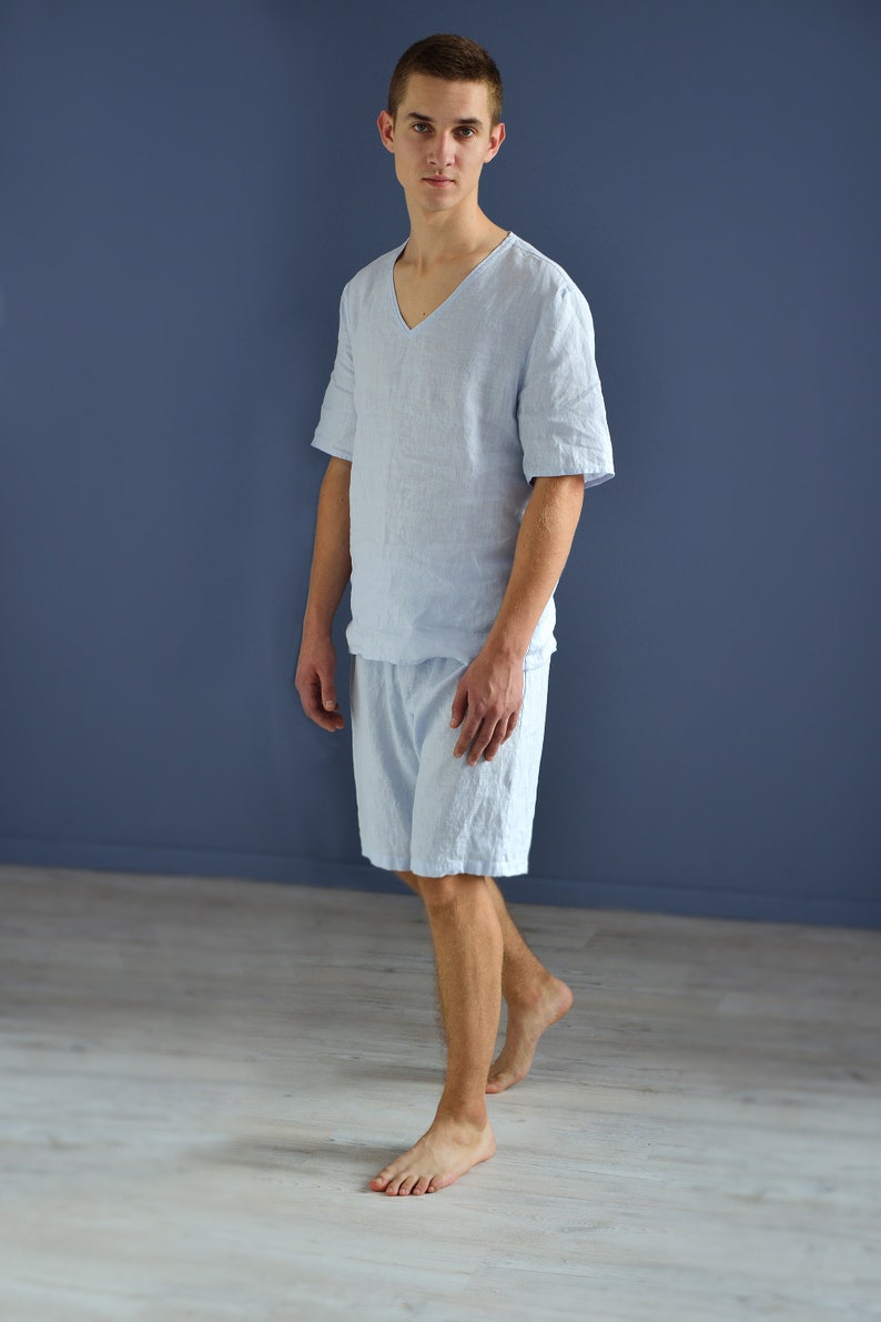 Pode incluir: Um homem vestindo uma camisa de linho azul claro e shorts combinando. Ele est&aacute; em p&eacute; em um piso de madeira clara em frente a uma parede azul.