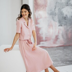 Puede incluir: Una mujer con un vestido de lino rosa con camisa de cuello y falda. Est&aacute; sentada en un cubo blanco frente a una pintura abstracta rosa y gris.