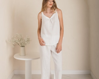 Peut inclure: Ensemble de vêtements blancs en lin composé d'un caraco et d'un pantalon court. Le haut a une encolure en V et un détail froncé au centre. Le pantalon est de style ample. La tenue est associée à des chaussures blanches. Une petite table blanche avec un vase de fleurs est en arrière-plan.