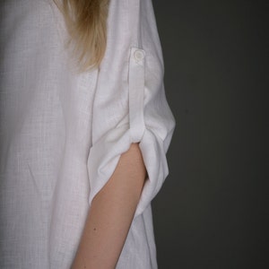 May include: A white linen shirt with a button on the cuff. The shirt is rolled up at the elbow.