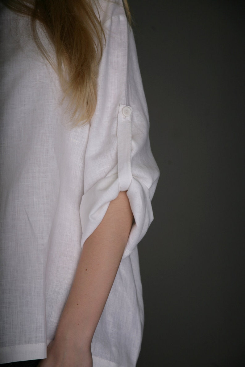 May include: A white linen shirt with a button on the cuff. The shirt is rolled up at the elbow.