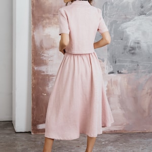 Puede incluir: Un vestido de lino rosa con un top corto y una falda. El vestido tiene un corpi&ntilde;o ajustado y una falda fluida. La modelo lleva el vestido con tacones blancos.