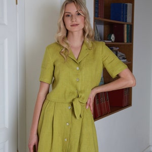 Puede incluir: Una mujer lleva un vestido de lino verde con botones en la parte delantera y un lazo en la cintura. El vestido tiene mangas cortas y una falda que le llega a las rodillas.
