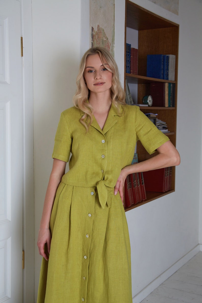 Puede incluir: Una mujer lleva un vestido de lino verde con botones en la parte delantera y un lazo en la cintura. El vestido tiene mangas cortas y una falda que le llega a las rodillas.