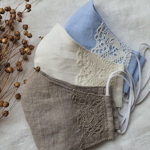 May include: Three linen face masks with lace trim. The masks are in shades of brown, white, and light blue.