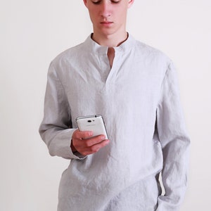Puede incluir: Un hombre con una camisa de lino gris claro con cuello mandar&iacute;n y mangas largas. Est&aacute; sosteniendo un tel&eacute;fono inteligente blanco en su mano derecha.