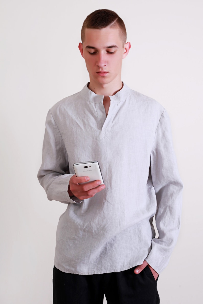 Puede incluir: Un hombre con una camisa de lino gris claro con cuello mandar&iacute;n y mangas largas. Est&aacute; sosteniendo un tel&eacute;fono inteligente blanco en su mano derecha.