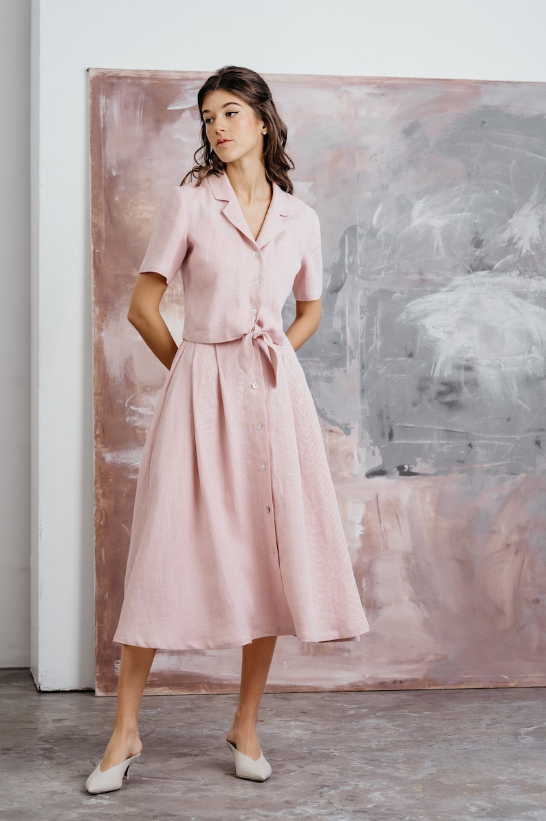 Puede incluir: Un vestido de lino rosa con cierre de botones en la parte delantera y un lazo en la cintura. El vestido tiene mangas cortas y una longitud midi.