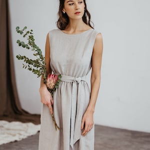 May include: A woman wearing a light gray linen dress with a self-tie belt. She is holding a bouquet of eucalyptus and a pink protea flower.