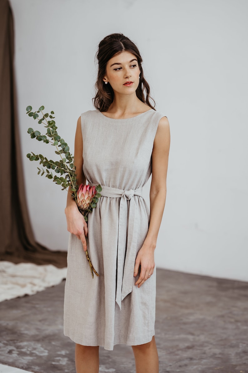 May include: A woman wearing a light gray linen dress with a self-tie belt. She is holding a bouquet of eucalyptus and a pink protea flower.