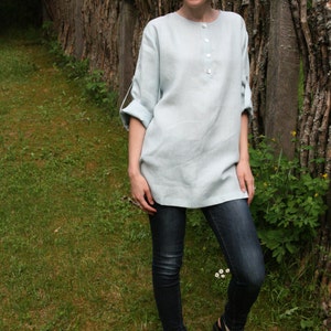 May include: A woman wearing a light blue linen shirt with a button-up front and jeans. She is standing in front of a wooden fence.