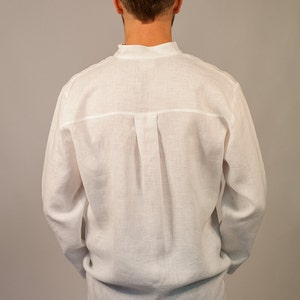 Puede incluir: Una camisa blanca de lino de manga larga y cuello abotonado. La camisa tiene un solo pliegue en la espalda.