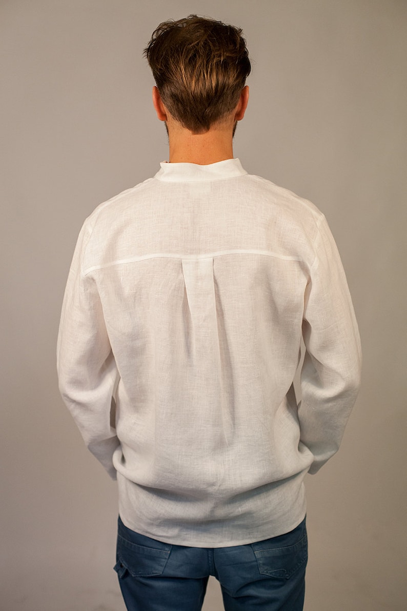Puede incluir: Una camisa blanca de lino de manga larga y cuello abotonado. La camisa tiene un solo pliegue en la espalda.