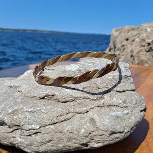 Könnte beinhalten: Ein geflochtenes Metallarmband mit abwechselnden Bändern in Gold-, Kupfer- und Silbertönen. Das Armband liegt auf einem grauen Stein, mit blauem Himmel und Wasser im Hintergrund. Das Design des Armbands ist eine durchgehende Drehung.