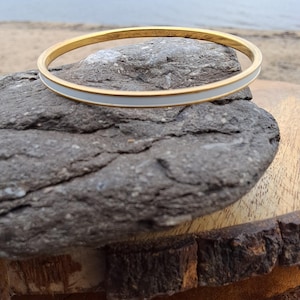 May include: A gold-tone bangle bracelet with a white enamel stripe. The bracelet is displayed on a gray rock with a blurred beach and ocean background. The jewelry is simple and elegant.